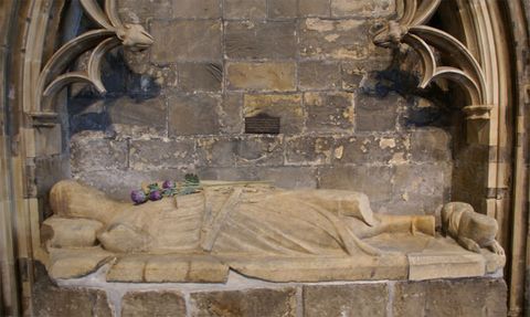 Sculpture. A carved effigy shows a man wearing robes lying on his back on a slab set in an arched niche decorated with tracery. One leg is broken off below the thigh.